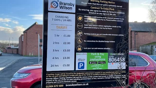 Welcome sign for Abbey Yard Pay & Display Car Park, detailing charges and contact information, with a parked car nearby.