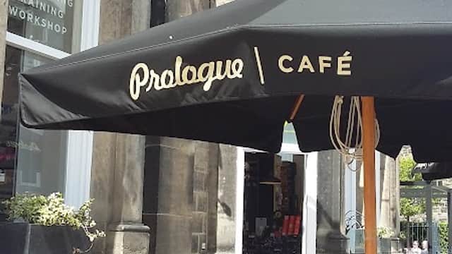 A black umbrella with "Prologue Café" text, outdoor seating, and a stone building with bike accessories displayed.