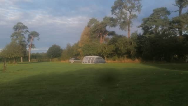 A spacious grassy campsite with a tent and a parked car, surrounded by trees under a partly cloudy sky.