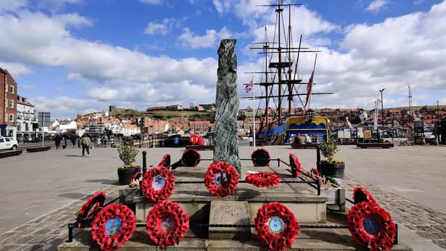 War memorial adorned with red poppies, overlooking a harbor with ships and a backdrop of Whitby's buildings and hills.