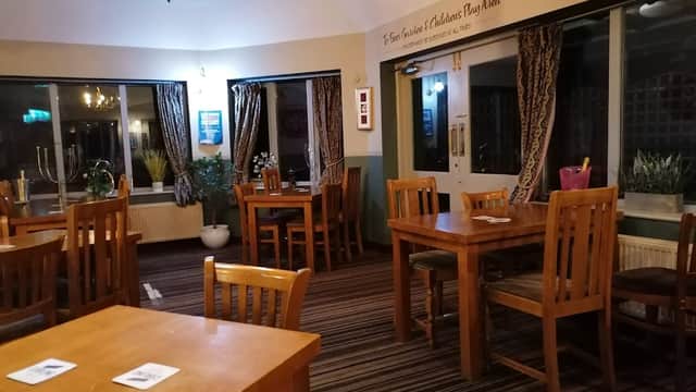 Cozy bar interior with wooden tables, chairs, patterned curtains, and potted plants in a warm, inviting atmosphere.