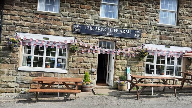 ARNCLIFFE ARMS B&B LTD sign, stone building, outdoor seating, flower pots, flags, and open door at 16:00.