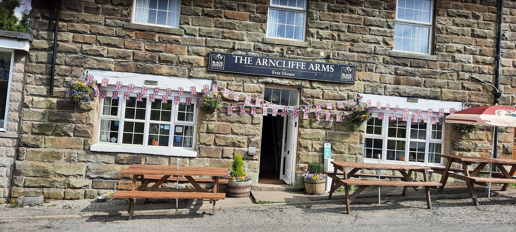 ARNCLIFFE ARMS B&B LTD sign, stone building, outdoor seating, flower pots, flags, and open door at 16:00.