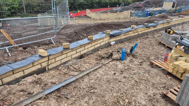 Construction site with a curved stone wall, gravel, and blue pipes, surrounded by fencing and building materials.