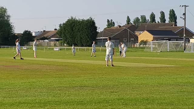 Cricket match in progress on a green field with players in white uniforms and houses in the background.