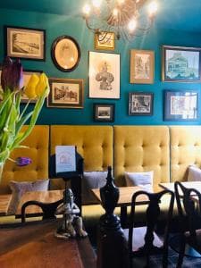 Cozy cafe with yellow tufted seating, teal walls, eclectic framed art, and a vase of tulips on wooden tables.