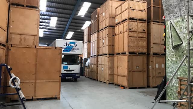 Wooden storage crates stacked in a warehouse with a removal truck labeled "Acomb" in the background.