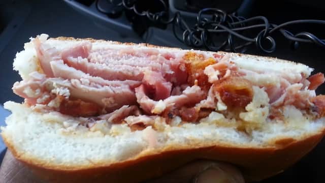 Close-up of a sandwich filled with layers of ham and crispy bacon, held in a hand, with a vehicle interior in the background.