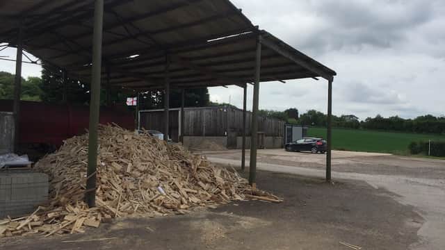 Woodpile under a metal roof, with a gravel area, buildings in the background, and a car parked nearby.