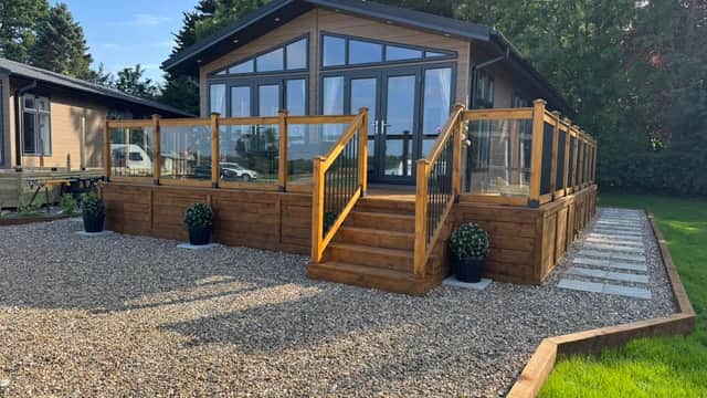 Modern lodge with wooden deck, glass railings, gravel pathway, and planters in a lush green setting.