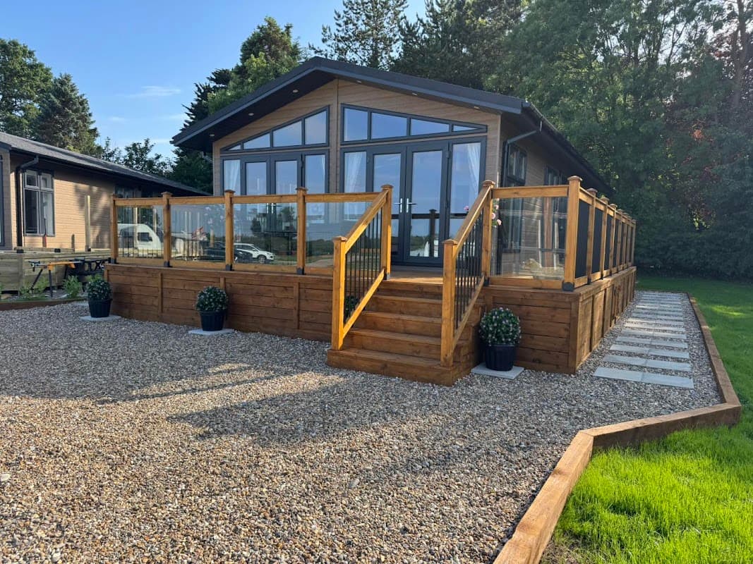 Modern lodge with wooden deck, glass railings, gravel pathway, and planters in a lush green setting.