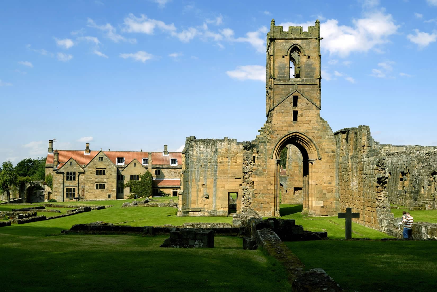 Ruins of Mount Grace Priory with a stone tower, grassy areas, and remnants of walls under a blue sky.