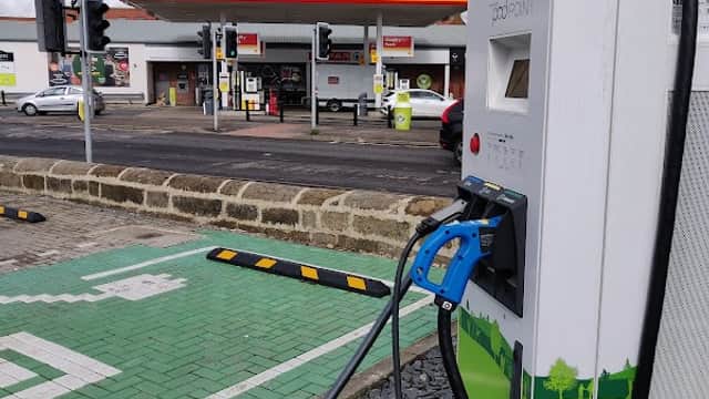 Pod Point charging station with cables, green charging bay, and a petrol station in the background under a cloudy sky.