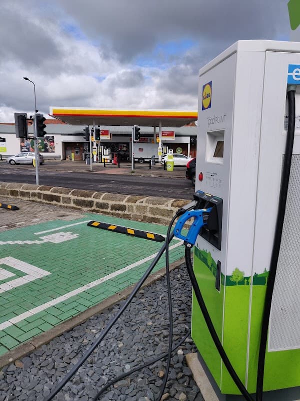 Pod Point charging station with cables, green charging bay, and a petrol station in the background under a cloudy sky.