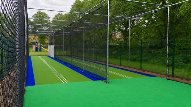 Cricket practice nets with green turf, surrounded by trees and a playground in the background.