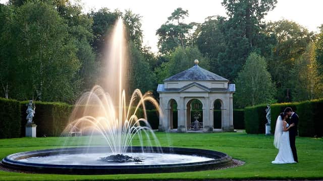 A couple embraces near a fountain in a landscaped garden, with a classical pavilion and trees in the background.