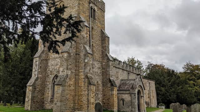 Historic stone church with a tall tower, surrounded by greenery and gravestones under a cloudy sky.
