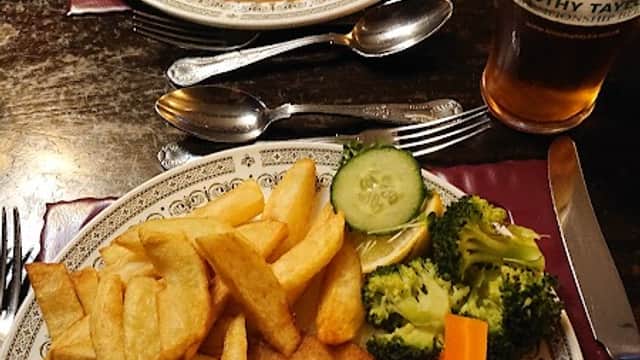 Two plates of hearty pub food with chips, vegetables, and a pint of beer on a wooden table in a cozy cafe setting.