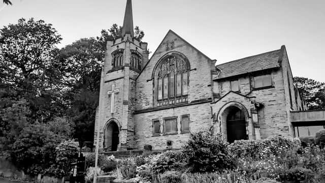 Historic stone church with a tall spire, surrounded by trees and flowering plants, in a quiet setting.