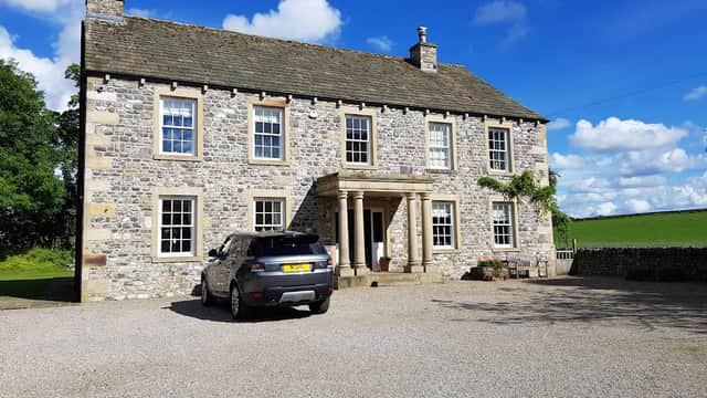 Stone building with a grand entrance, surrounded by greenery and a gravel driveway, featuring a parked car in front.