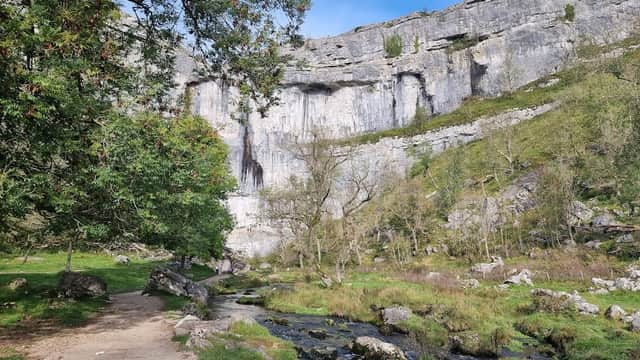 Lush green landscape with a stream, towering limestone cliffs, and a clear blue sky at Malham Cove, Yorkshire.