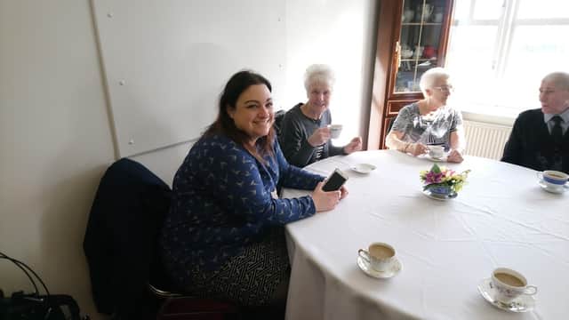 A group of four people enjoying tea at a round table in a bright room, with a floral centerpiece.