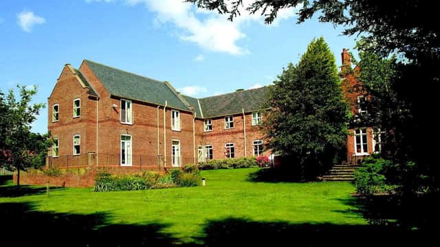 Red-brick manor house surrounded by lush green lawns and trees under a blue sky with scattered clouds.