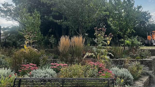 Park scene featuring a black bench surrounded by colorful flowers, shrubs, and lush greenery under a cloudy sky.