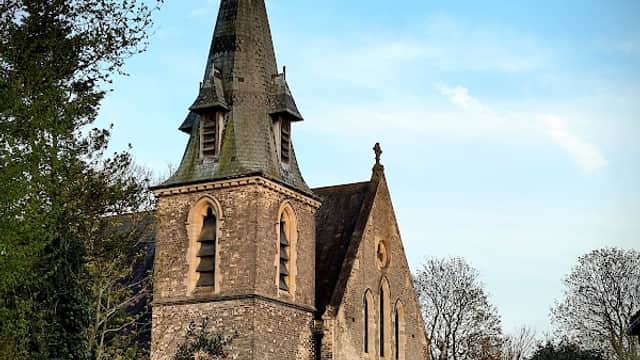 Stone church with a tall spire, surrounded by trees and a well-kept lawn under a clear blue sky.