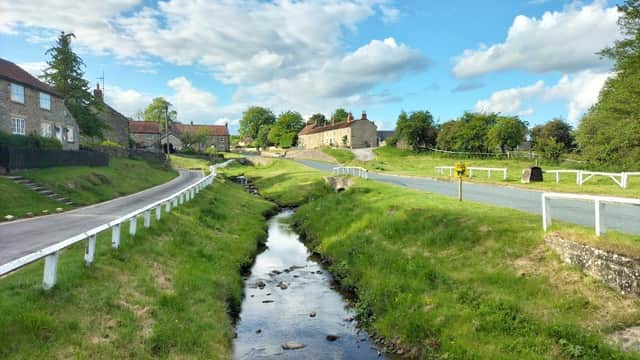 Scenic view of Hutton-Le-Hole Caravan Site with green fields, a stream, and charming cottages under a blue sky.