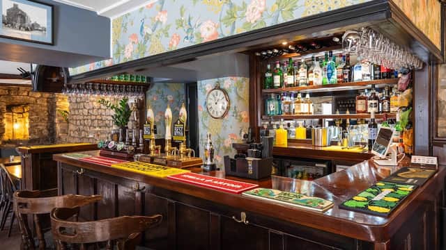 Wooden bar with floral wallpaper, shelves of drinks, clocks, and bar stools in a cozy pub setting.