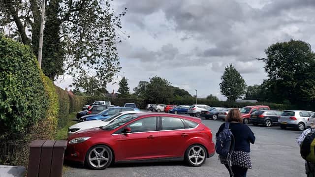 A busy car park featuring various parked cars, with a red car in the foreground and trees lining the background.