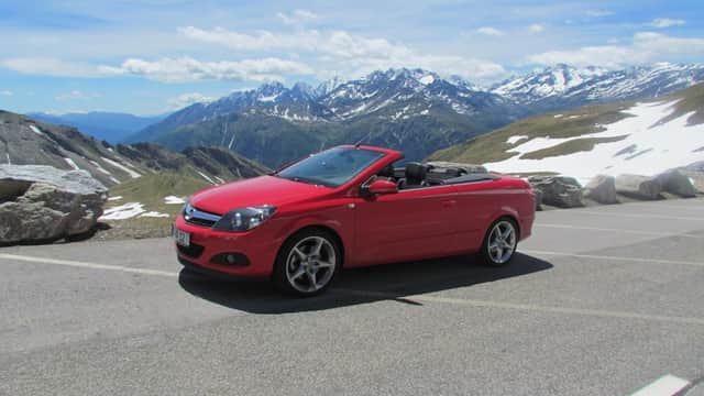 Red convertible car parked on a mountain road with snow-capped peaks and blue sky in the background.
