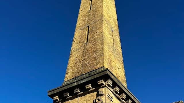Tall stone tower against a clear blue sky, part of Salts Mill with nearby historic buildings. Free parking area visible.