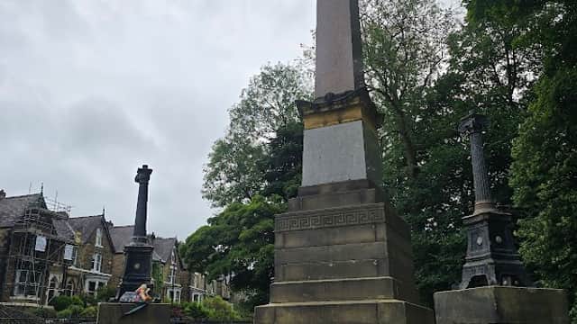 Tall stone obelisk surrounded by greenery, with ornate lampposts nearby and residential buildings in the background.