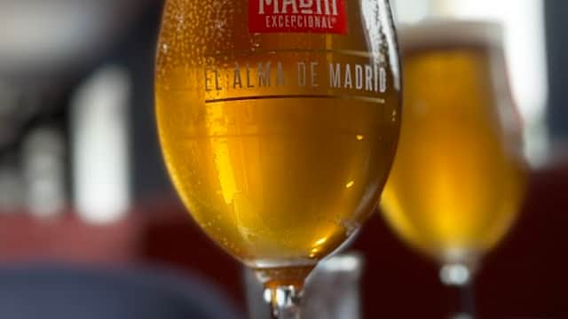 A close-up of a frosty beer glass with a logo, sitting on a wooden table, with a blurred background of another glass.