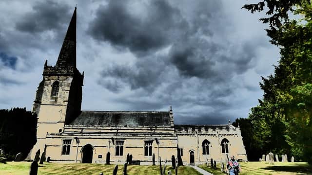 St Peter's Church with a tall spire, surrounded by gravestones and lush greenery under a dramatic cloudy sky.