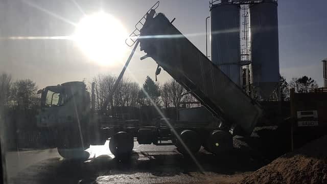 A truck unloading material beside large silos, with sunlight shining in the background and a sandy pile nearby.