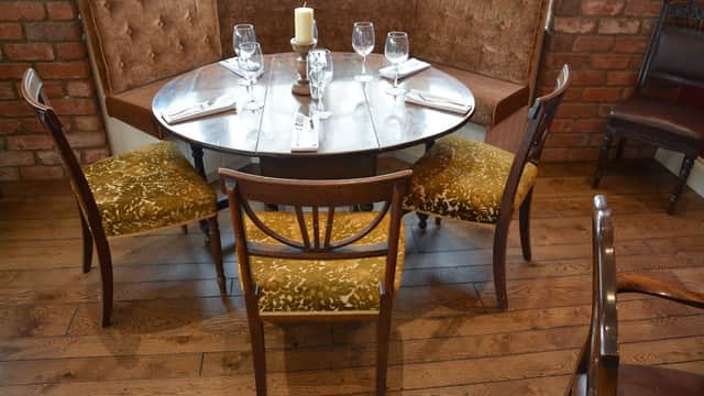 Cozy dining area with a round wooden table, elegant glassware, and upholstered chairs against a brick wall.