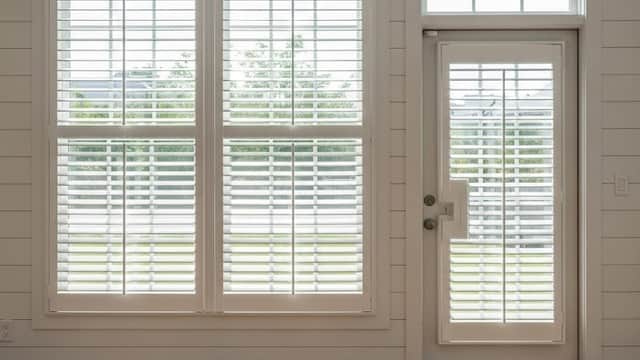 Bright interior featuring white window shutters on large windows and a glass door, with natural light illuminating the space.