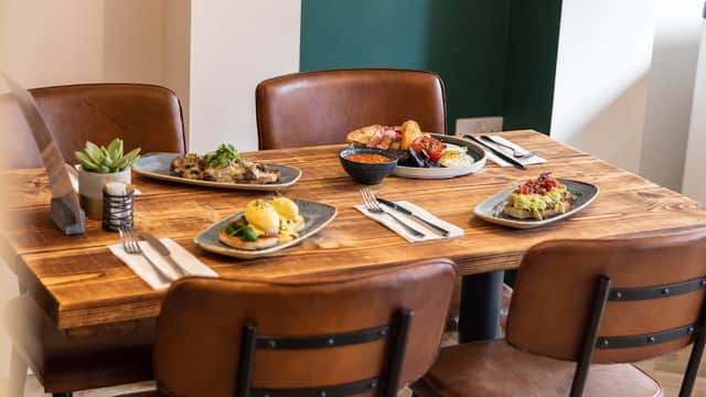 A wooden table set with five plates of food, cutlery, and a small plant in a pot, surrounded by leather chairs.