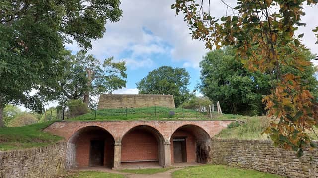 Arched brick entrance leading to a grassy mound under a cloudy sky, surrounded by trees and greenery.