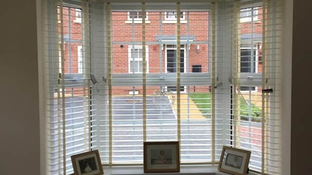 Bay window with white blinds, framed pictures on the sill, and a view of a brick building and garden outside.