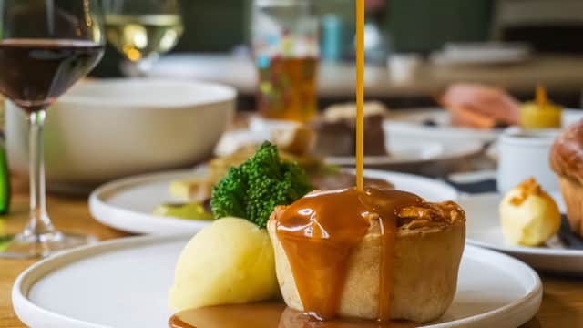 A plate with a meat pie drizzled in gravy, alongside mashed potatoes and broccoli, with wine glasses in the background.