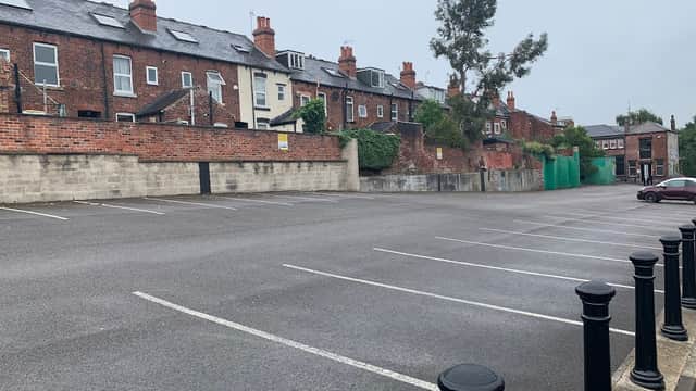 Empty car park with brick buildings and chimneys in the background, surrounded by greenery and a cloudy sky.
