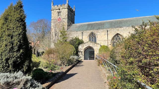 Historic church with a tower, surrounded by lush greenery and colorful flowers under a clear blue sky.