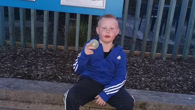 A child in a blue jacket sits on a bench holding a ball, with a signboard and wooden fence in the background.