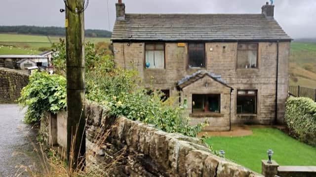 Stone building with a slate roof, surrounded by greenery and a dry stone wall, under a cloudy sky.