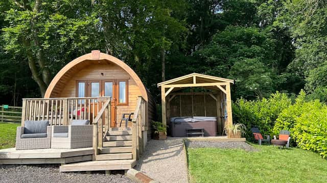 Wooden glamping pod with steps, surrounded by greenery, and a gazebo with a hot tub nearby on a gravel path.