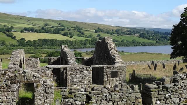 Ruins of stone structures with green hills and Semer Water lake in the background under a partly cloudy sky.